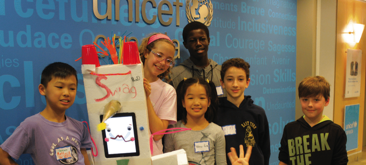 Un groupe diversifié de six enfants se tient ensemble, souriant, devant un fond bleu de l'UNICEF Canada. Ils tiennent un projet créatif fabriqué à partir de divers matériaux, avec un visage animé sur un écran et le mot 'Swag' écrit dessus. Le cadre suggère un atelier ou un événement axé sur l'engagement des jeunes.